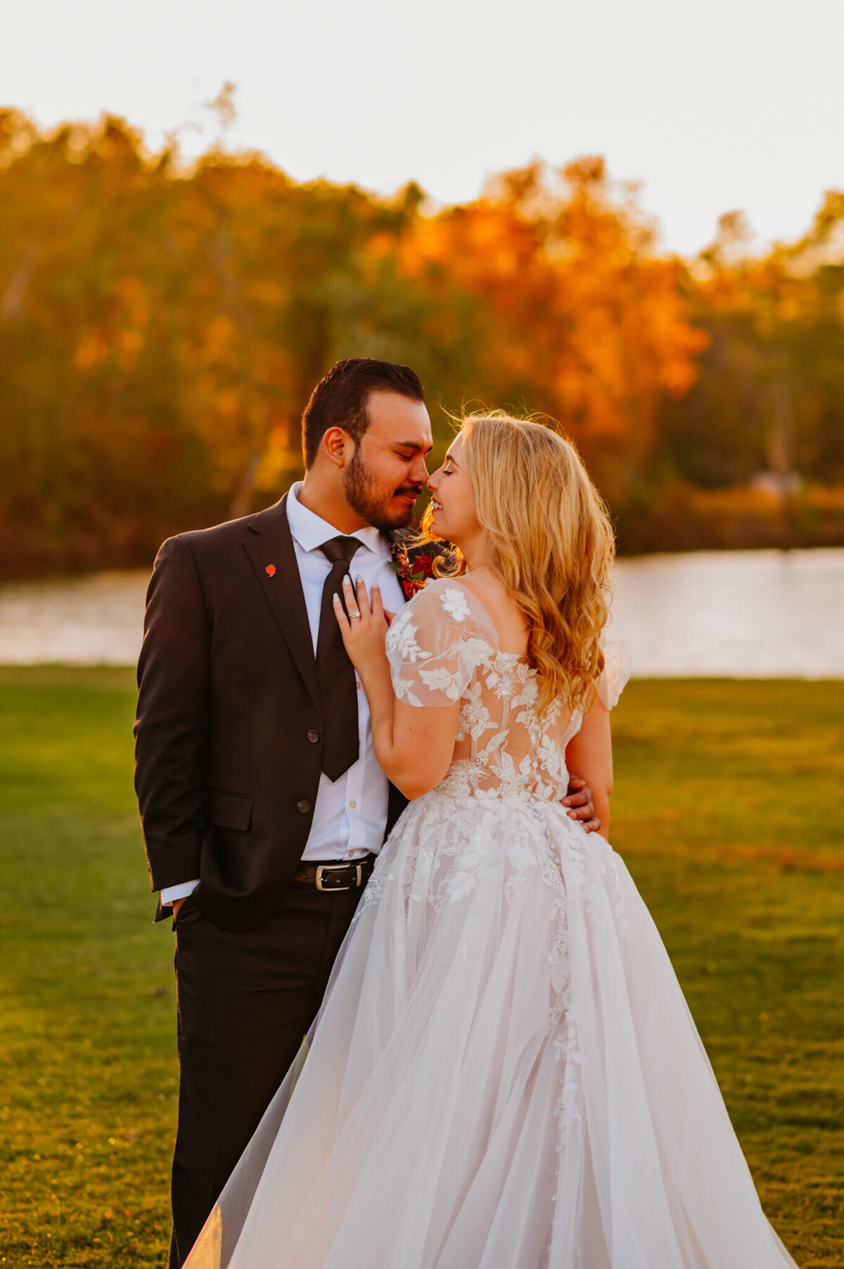 photo of a bride and groom rubbing their noses together with a pond, fall foliage, and the sun in the background