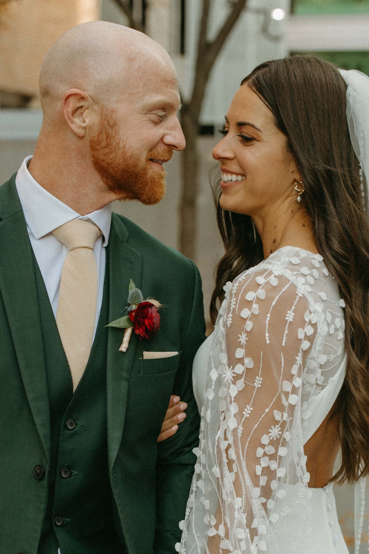 A bride and groom smiling at each other in a park, the bride in a detailed lace dress and the groom in a green suit with a red boutonniere.