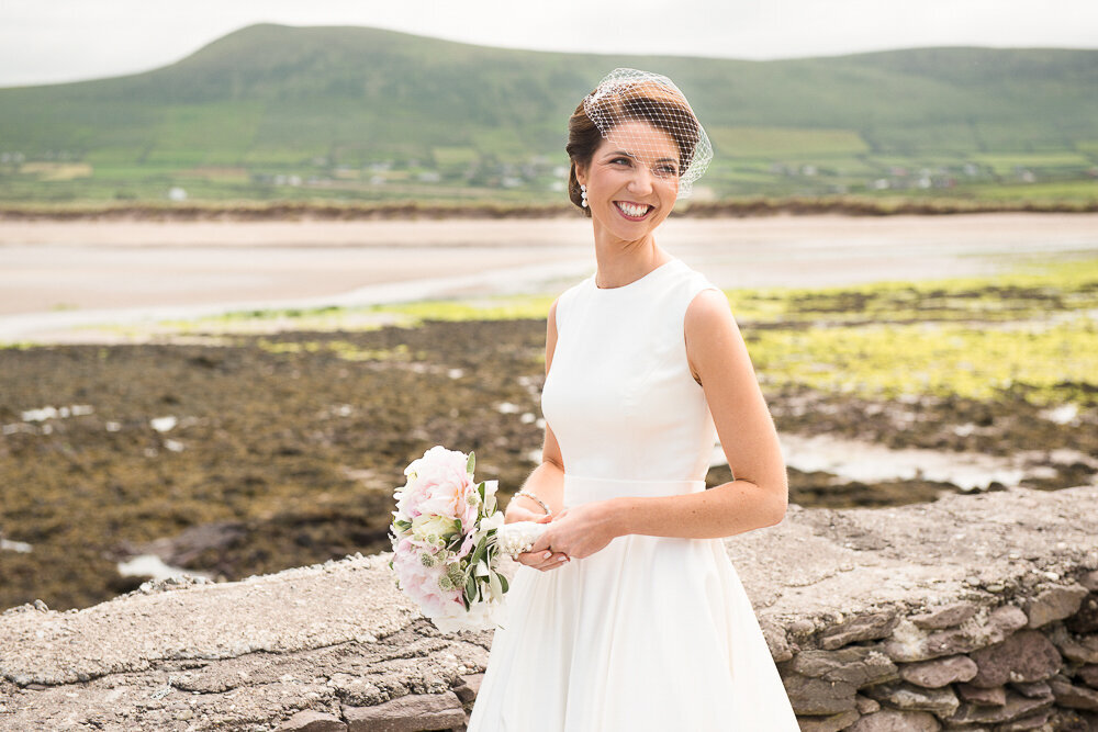 brunette bride wearing a birdcage veil and tea-length, vintage wedding dress, holding a bouquet with light pink roses while standing by a stone wall with the sea and mountains in the background