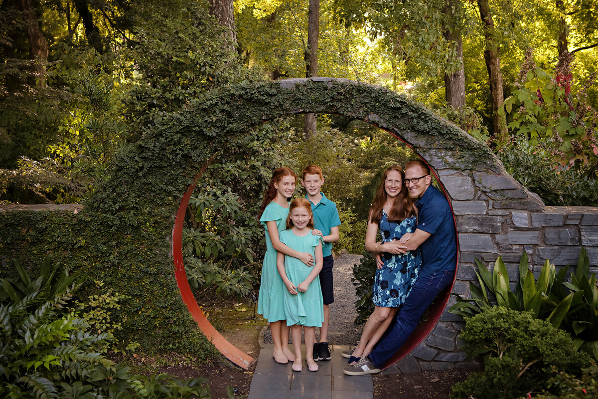 Spring family portrait at UNCC Botanical garden in the Moongate concrete circular archway. 3 kids plus mom and dad who are hugging and leaning against the curvature of the moongate structure. 