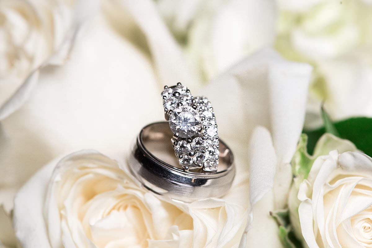 The bride's diamond engagement ring and wedding band are sitting on top of the groom's silver wedding band. They are nestled into a bed of white roses.