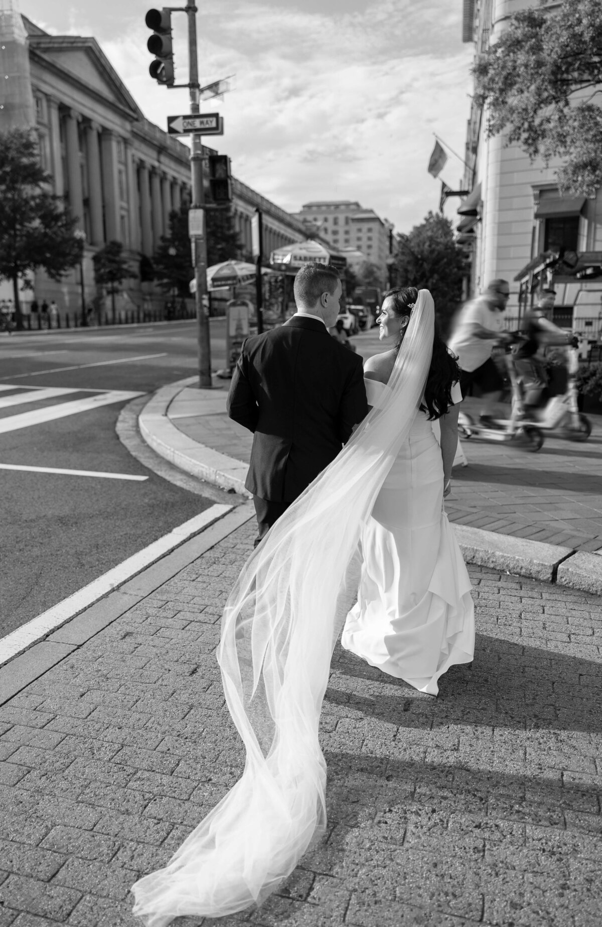 A bride and groom walk hand in hand on a city street. The brides long veil flows behind her. The scene is in black and white, capturing a busy urban setting with buildings and pedestrians.