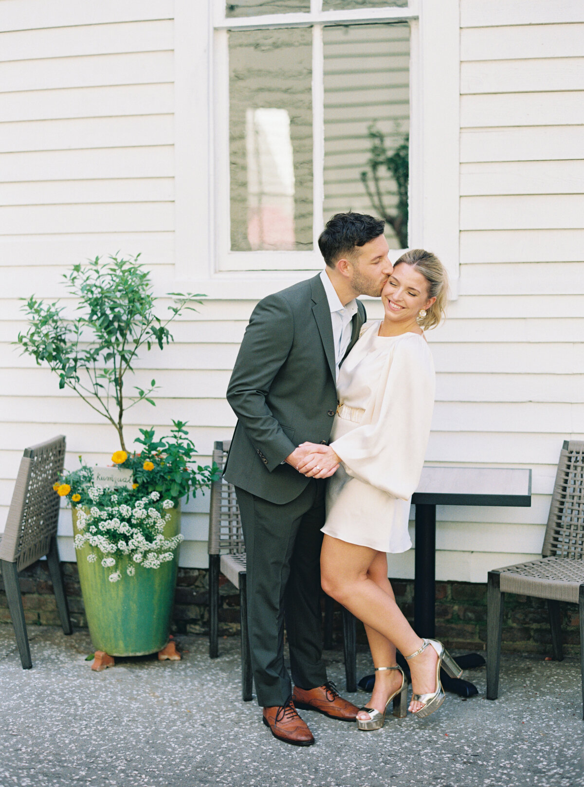 Guy in olive green suit kissing girl in short white dress with flowy sleeves. Film engagement session in downtown Charleston.
