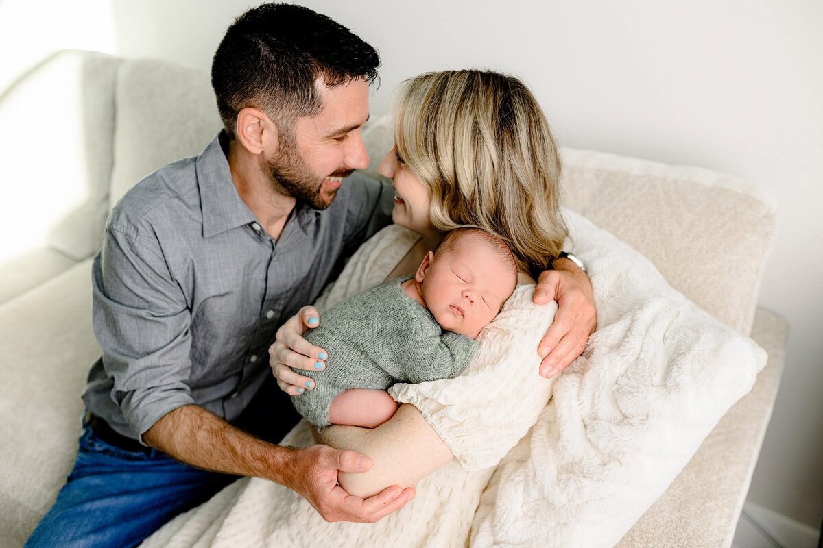 mom and dad look at each other while sitting on their sofa and mom is holding her son on her shoulder