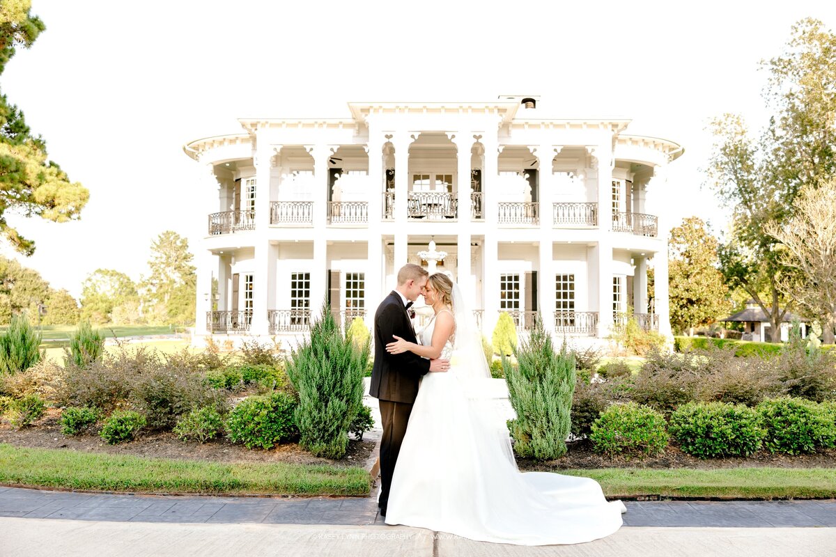 A bride in a white gown and groom in a black tuxedo embrace in front of a grand, white-columned mansion, an elegant setting for luxury wedding planning.