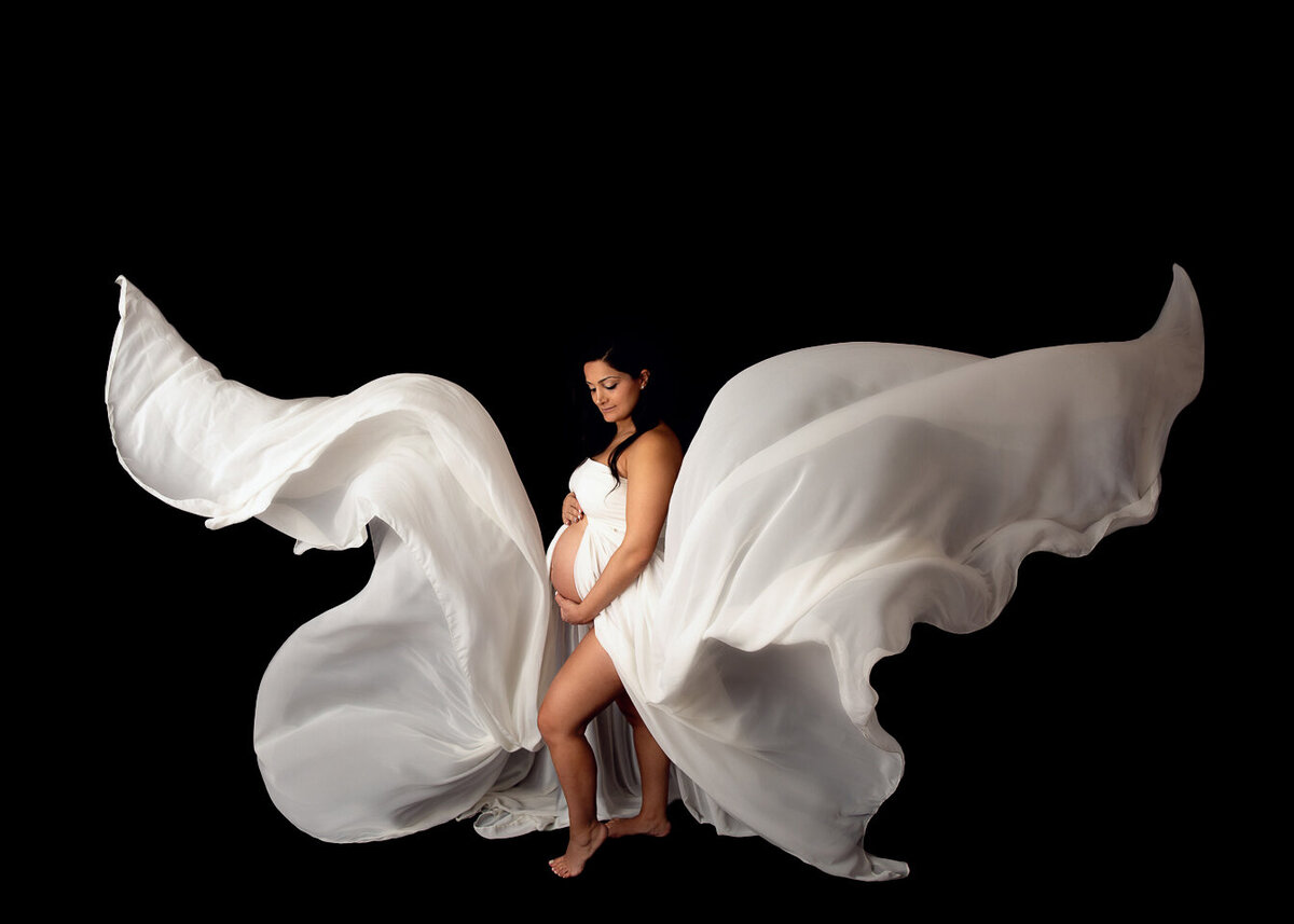 A mother to be covering with a white sheet stands in a studio as the ends blow in the wind in a New Jersey Maternity Photographer studio