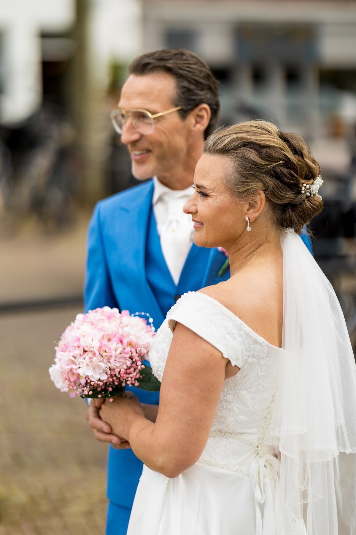 Bruidspaar van de zijkant gefotografeerd, met zichtbaar roze bruidsboeket, perfect georganiseerd door weddingplanner