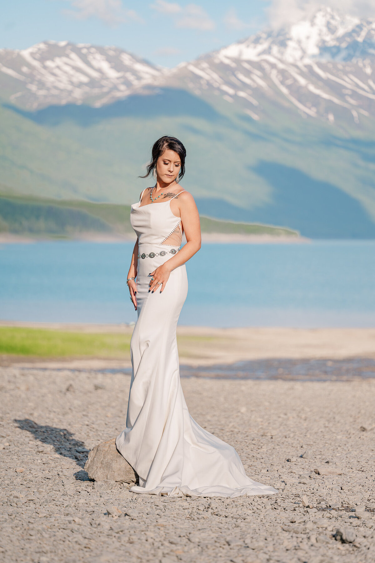 A brunette bride next to a lake and mountains during the midnight sun in the summer enjoying her Raleigh wedding photos