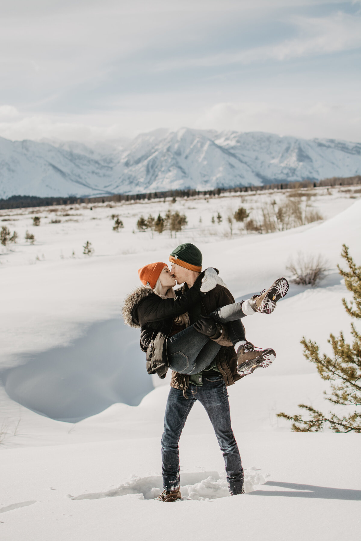 winter engagmeent session with jackson hole photographers capturing man holding his fiance and carrying her through the snowas they romantically kiss  for their winter engagement session