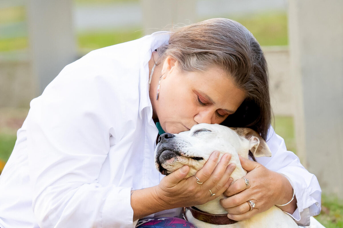 Woman hugging and kissing her elderly Pitbull dog by a white wooden fence