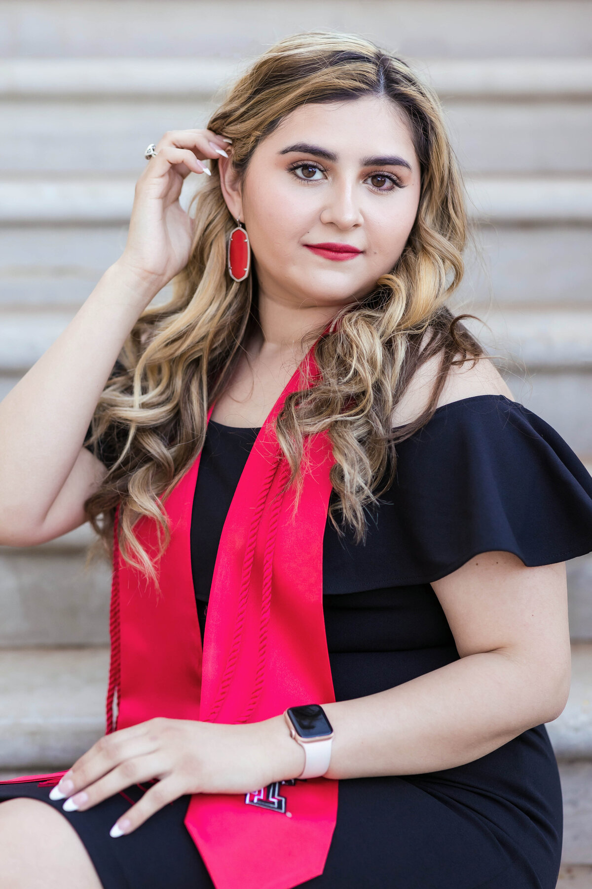 Hispanic female college graduate session on Texas Tech Campus.  Young woman is wearing a black dress with red college stole and red earrings and is holding her hair while looking at the camera