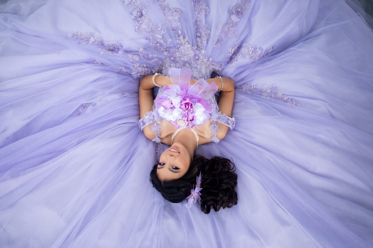 teenage girl lays on her purple ballgown holding purple and white bouquet Untermeyer Park New York Westchester quinceanera Photographer Milestone images
