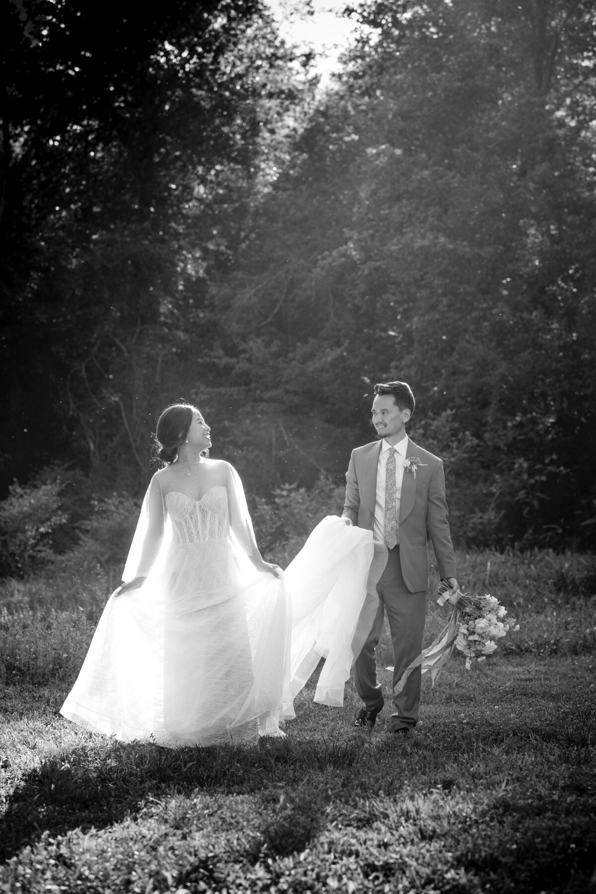 Asian-bride-and-groom-walking-in-rays-of-sunlight-smiling-while-the-groom-carries-the-bride's-train-at-the-Providence-Cotton-Mill-in-Charlotte-NC