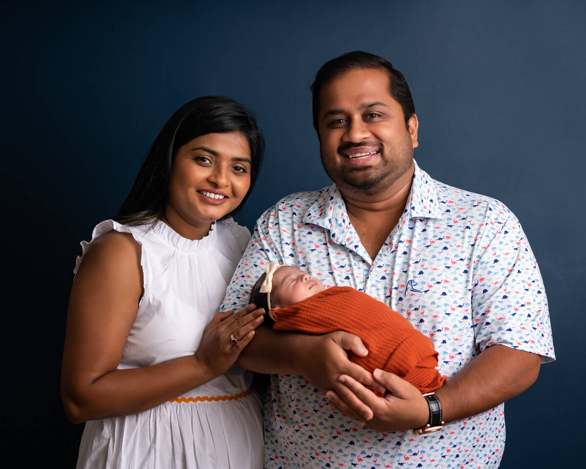 A mama and daddy snuggle their newborn baby girl who is wrapped in an orange swaddle
