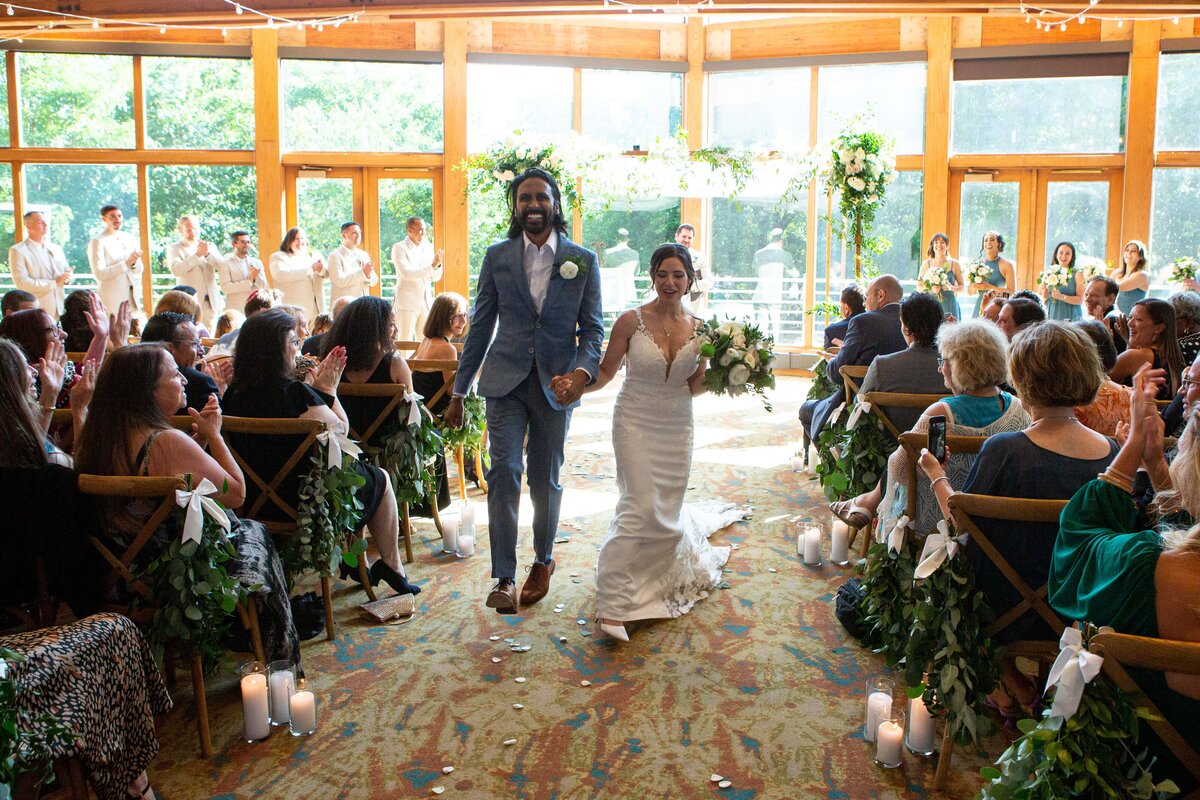 A bride and groom joyfully walking down the aisle after their wedding ceremony, orchestrated by a wedding planner in Des Moines, surrounded by guests in a sunlit room adorned with greenery.