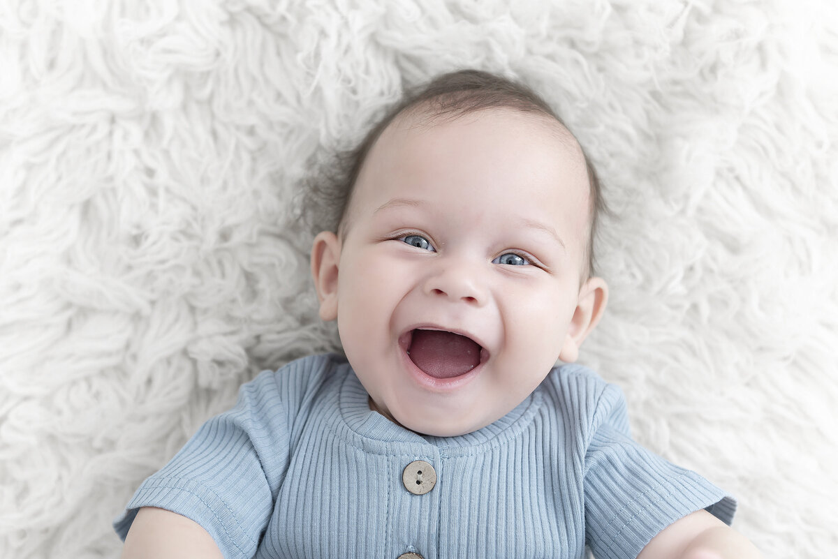 A smiling young infant boy lays on a shag blanket in a blue onesie