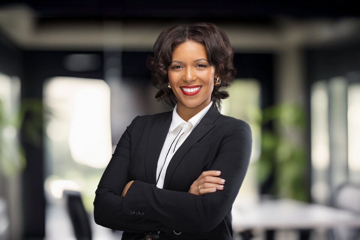 A professional African American woman with short, styled hair and a confident smile, dressed in a tailored black blazer and crisp white blouse. She stands with her arms crossed in a modern office setting, featuring blurred greenery and large windows in the background. Captured by a Cincinnati-based headshot photographer specializing in corporate and personal branding, this image exudes professionalism and approachability, making it ideal for LinkedIn profiles and marketing materials.