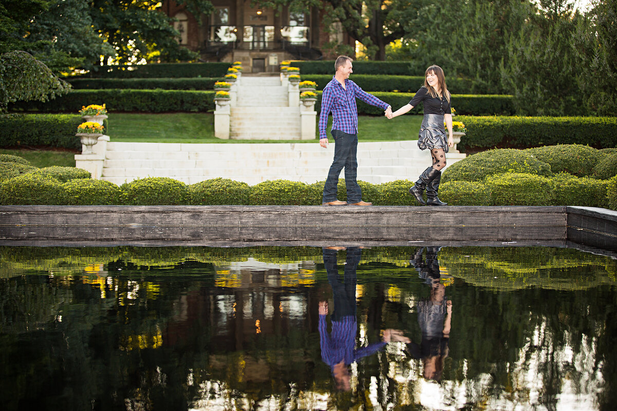 An engaged couple with casual outfits are walking near a pool with beautiful greenery  in Cantigny Park in Wheaton, IL