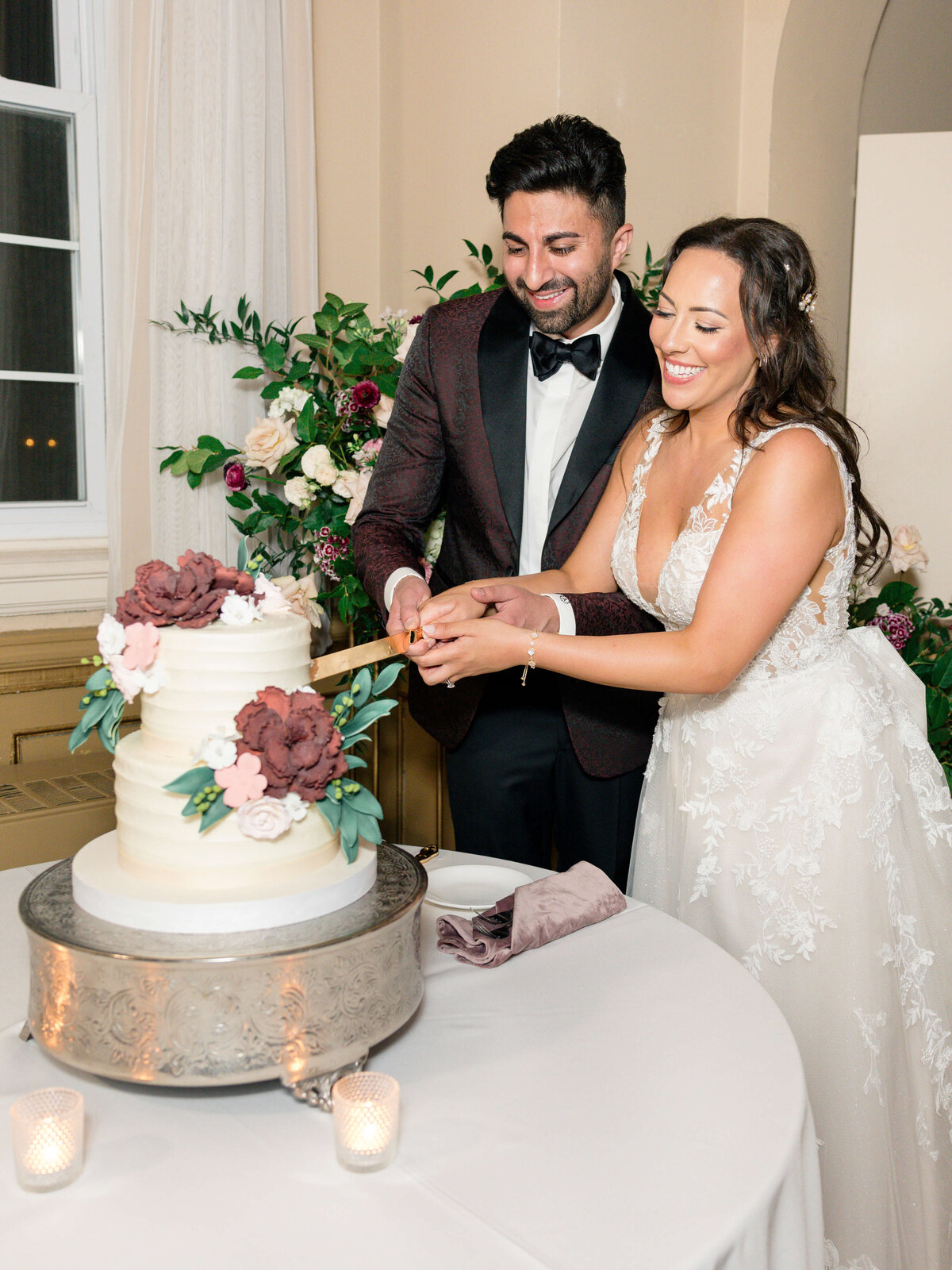 A bride and groom smile as they cut a tiered wedding cake adorned with burgundy and white flowers. The bride wears a white lace gown, and the groom is in a dark suit with a bow tie. The table is decorated with candles and flowers.