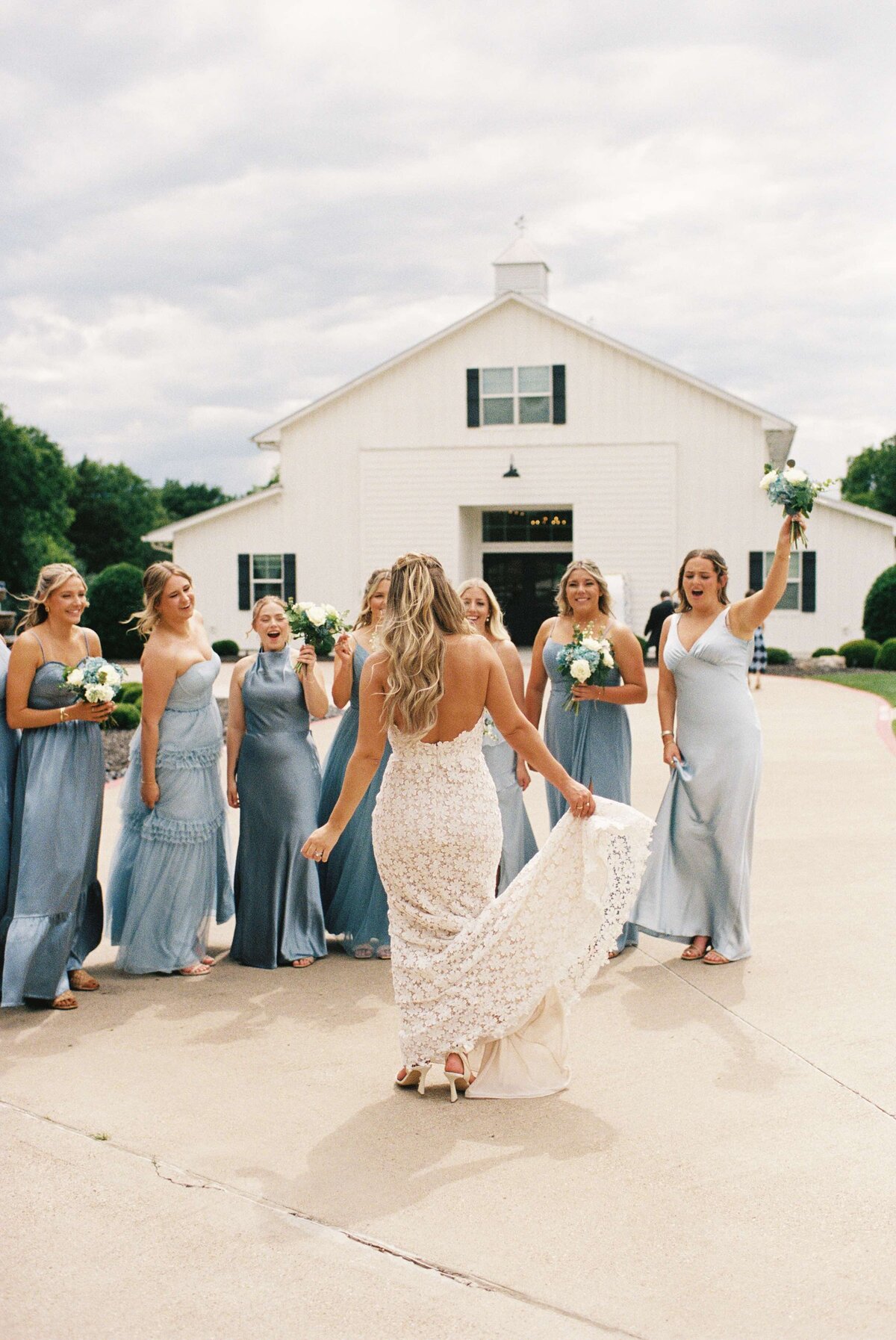 Bride in lace dress walks toward joyful bridesmaids in pale blue dresses holding bouquets. They're in front of a white barn on a cloudy day.