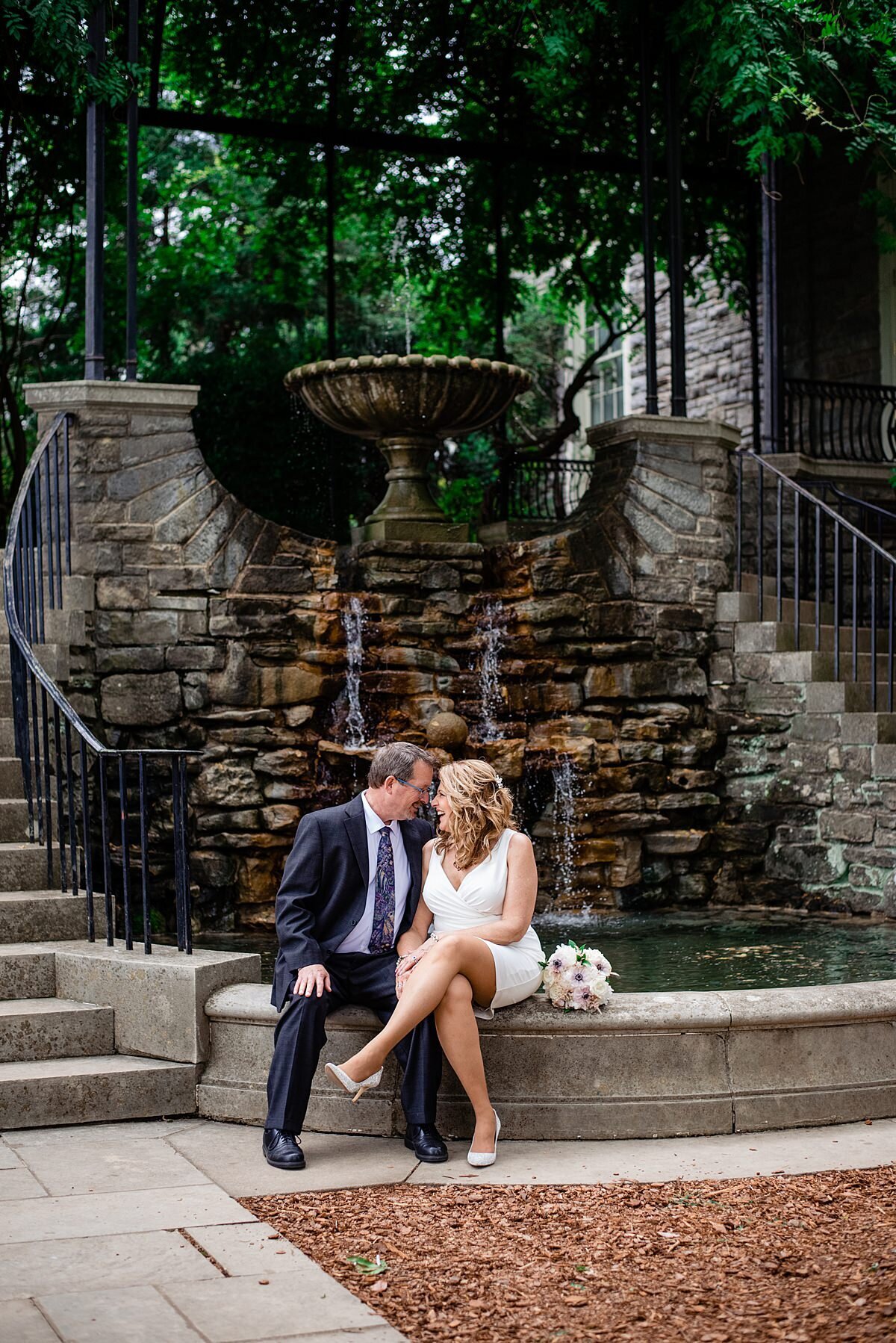 Sitting at the base of a waterfall  fountain with a large stone chalice at the top, the bride and groom touch foreheads as the groom puts his arm around the bride's waist. There are two semi spiraling staircases  on either side of the fountain. The groom is wearing a navy suit with a white shirt and blue patterned tie. The bride is wearing a short, fitted white dress with a plunging neckline. The bridal bouquet sits on the edge of the fountain  next to her. It is made up of white, ivory and blush flowers.