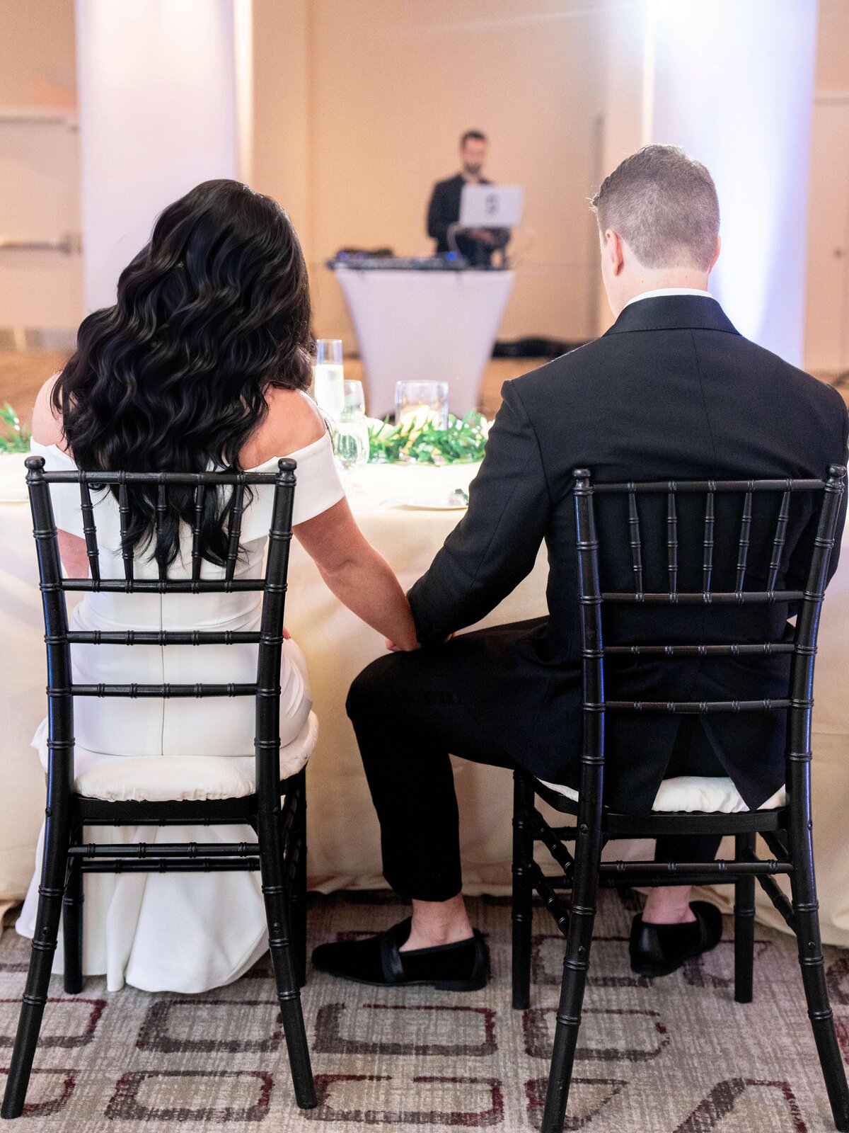 A couple wearing formal attire is seated at a table, holding hands and facing away. They are on black chairs with greenery on the table in front of them. A person with a laptop stands in the background.