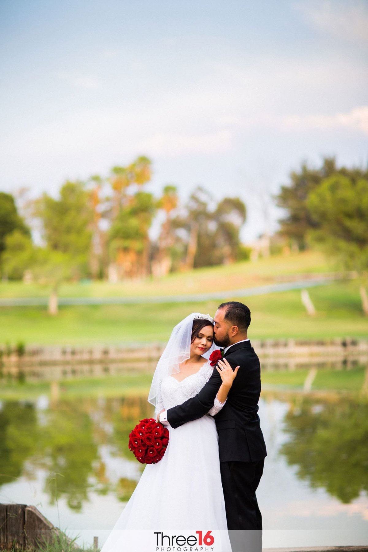 Groom embraces his Bride and kisses her forehead in front of a golf course lake