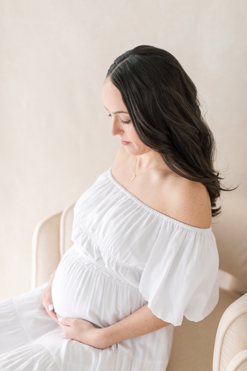 A pregnant woman wearing a white off the shoulder dress sits on a chair and holds her pregnant belly as she gazes down at it during her Lawrenceville NJ maternity photography sessions