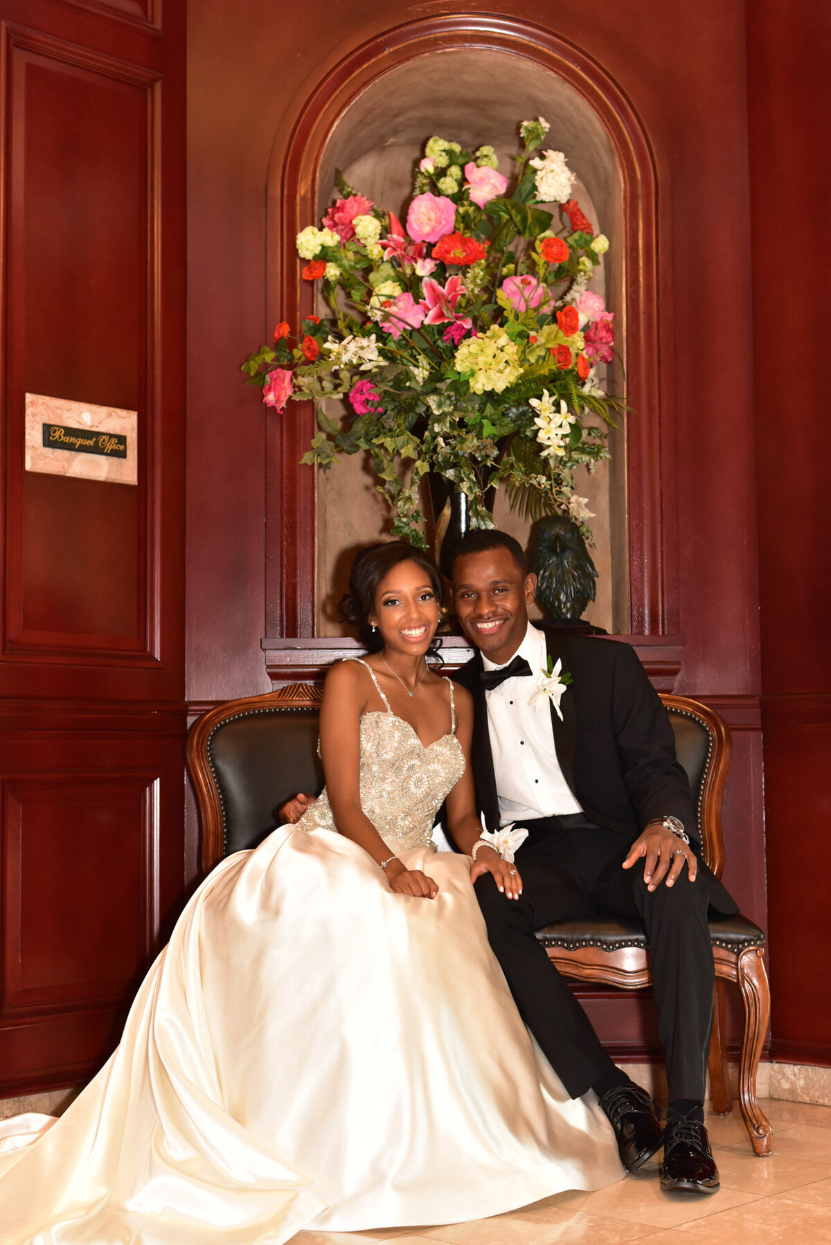 Bride and groom smiling while sitting down in a sofa that has florals behind