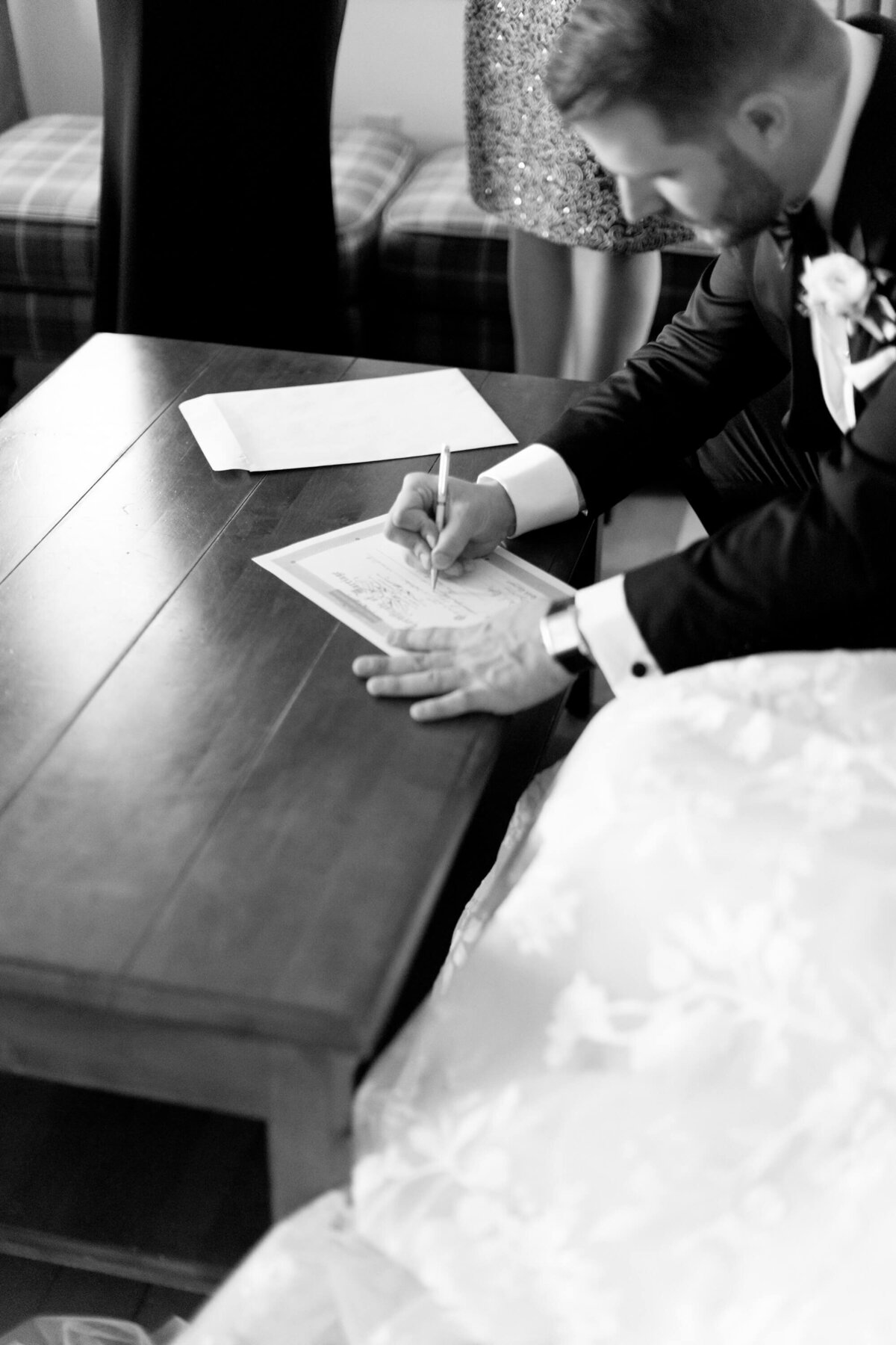 Handsome bridegroom-to-be in black wedding suit and a watch signs the marriage registration certificate on a tabletop.