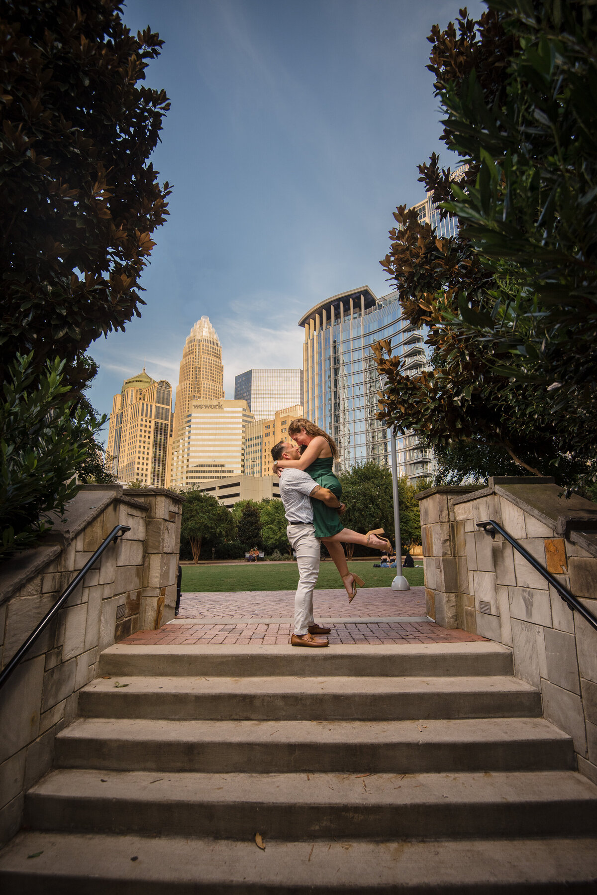 Man-lifting-his-fiancée-up-at-the-top-of-the-stairs-in-Romare-Bearden-Park-with-city-building-in-the-background