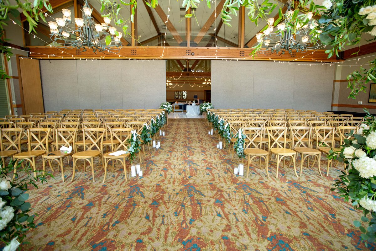 A wedding hall setup with rows of wooden chairs on both sides of a central aisle, decorated with floral arrangements and lanterns on the floor, leading to a decorated table at the front for an Iowa wedding