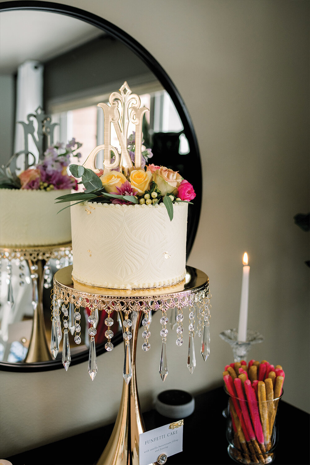 Elegant baby shower cake with lace icing details, adorned with pink and yellow flowers, and topped with a custom rose gold monogram acrylic topper, displayed on a rose gold and crystal cake stand.