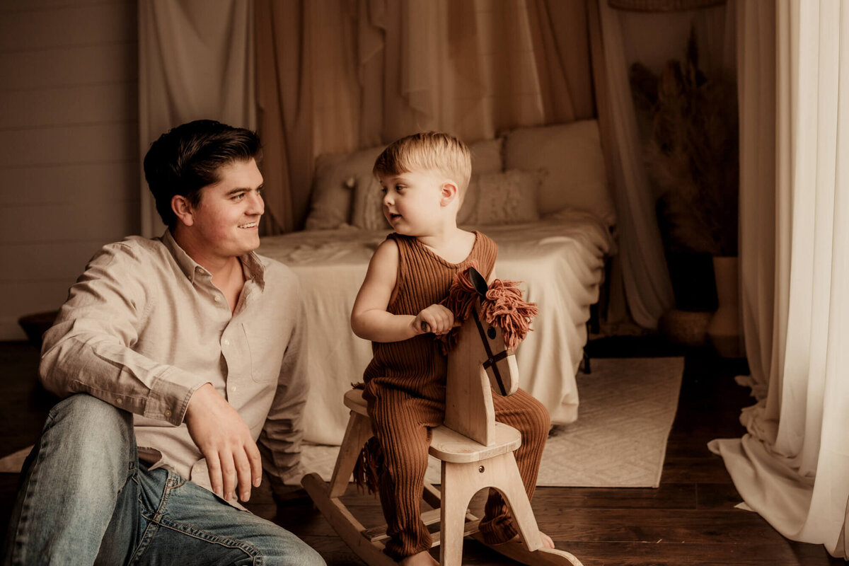 Father and son smiling at one another as the boy sits on a rocking horse.