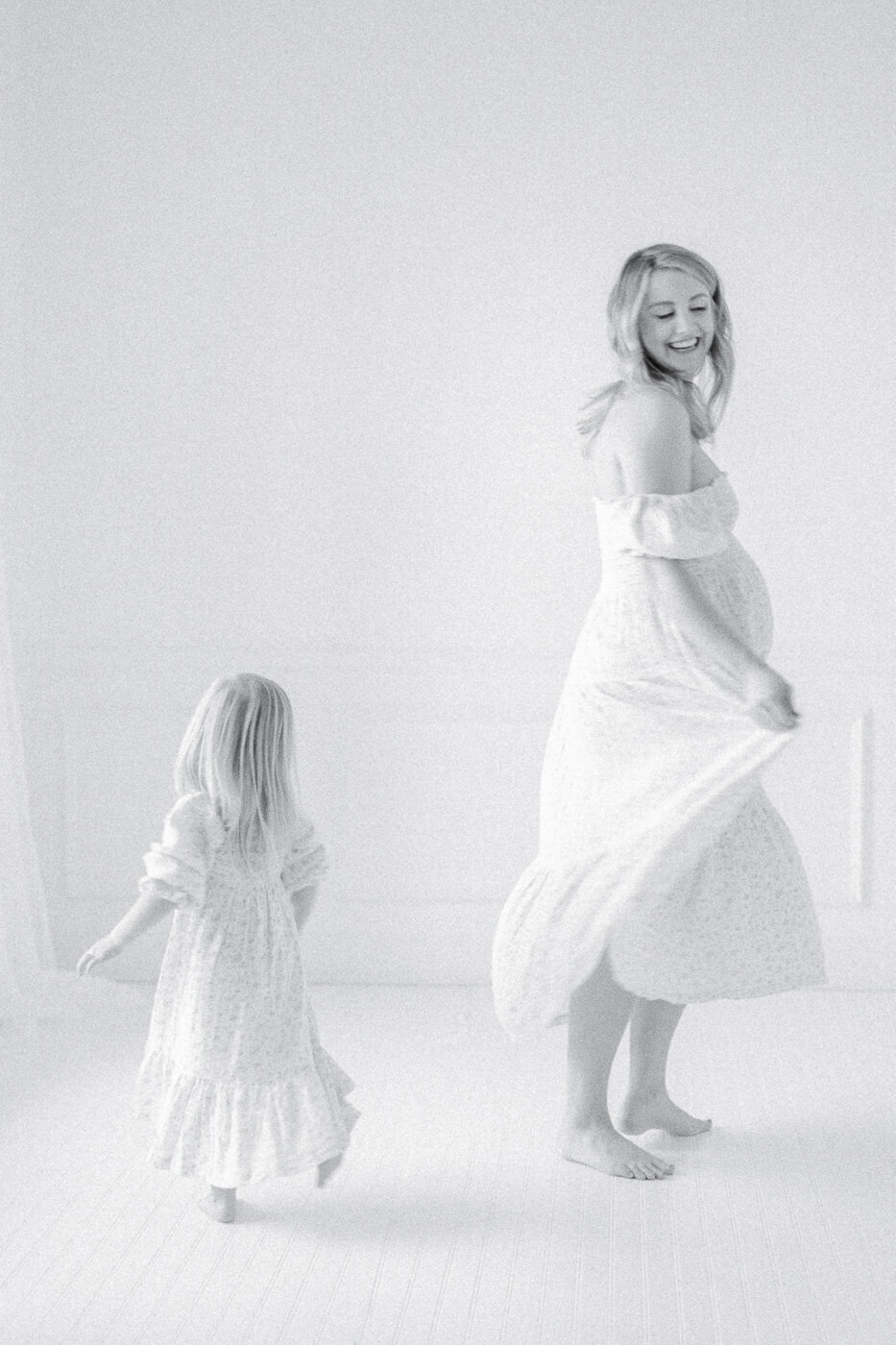 Black and white image of a pregnant mom and her daughter spinning in white dresses