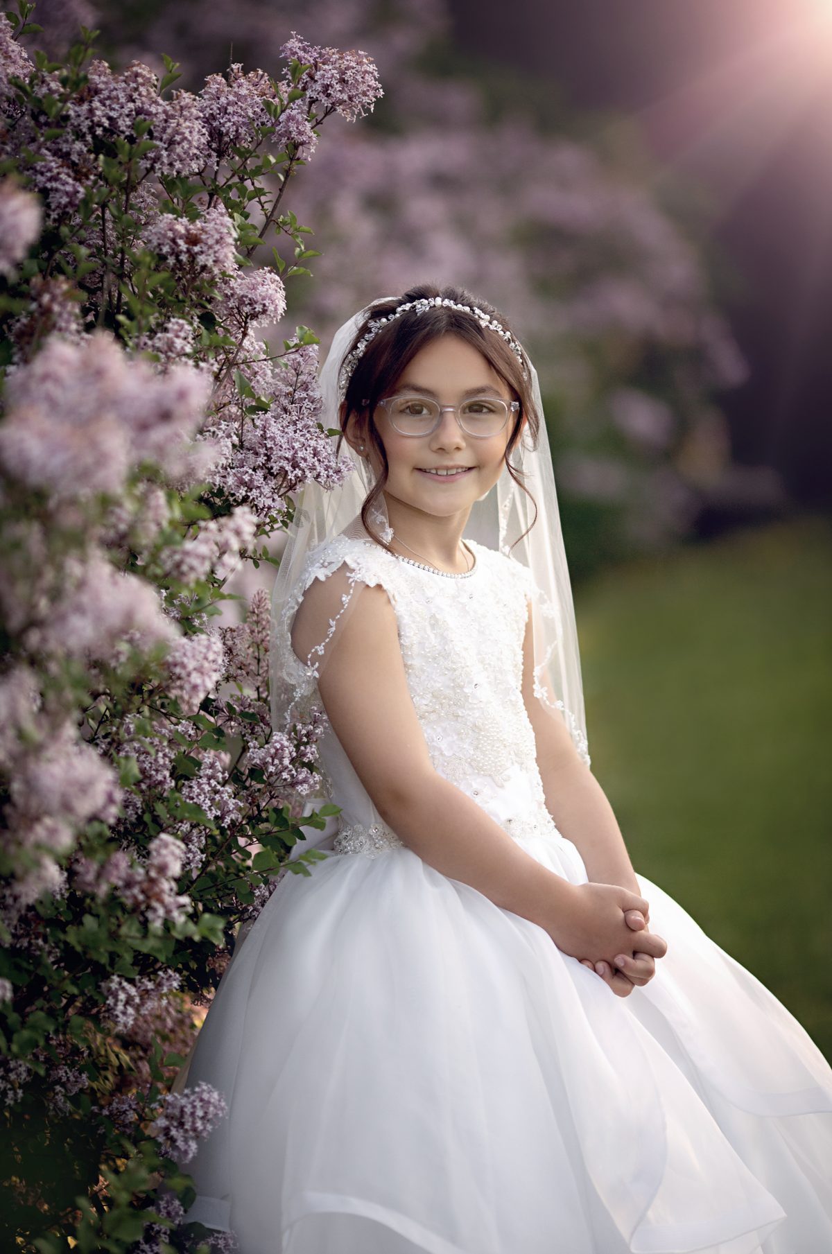 A young girl with glasses stands among some pink flowering bushes in a white communion dress