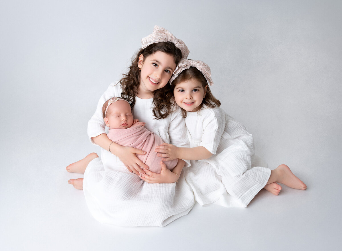 Two big sisters hold their new baby sister for a posed studio newborn portrait in Brooklyn, NY.