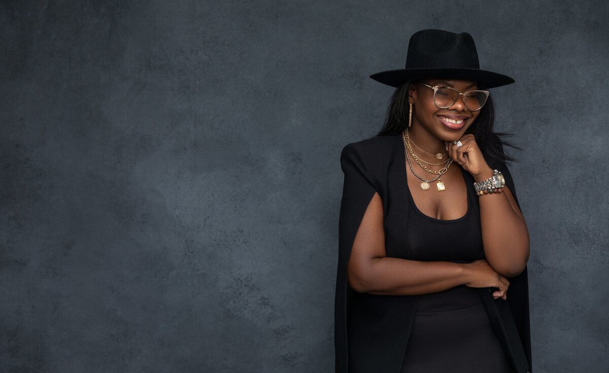 Striking Cincinnati portrait of a confident woman wearing glasses and a stylish black hat. Her radiant smile, layered necklaces, and dark attire create a captivating and modern look.