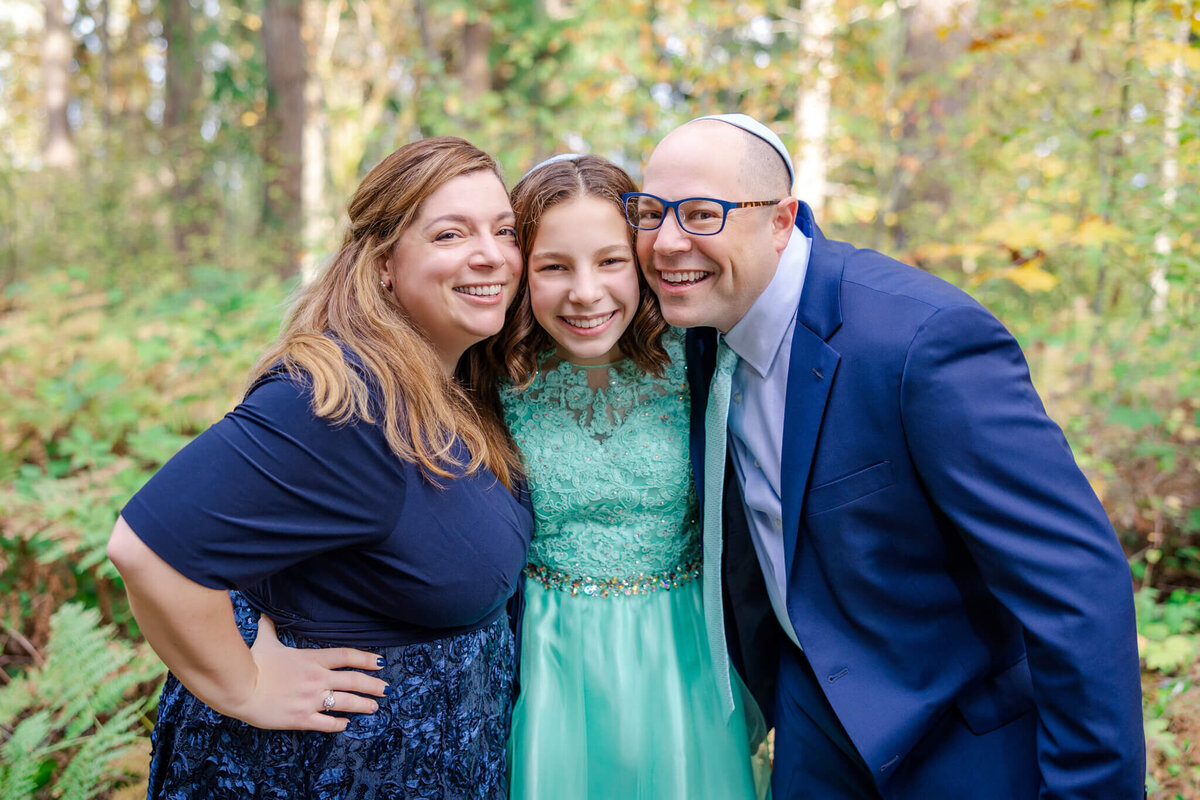 A proud mom and dad hug onto their teen daughter in a green dress in a forest