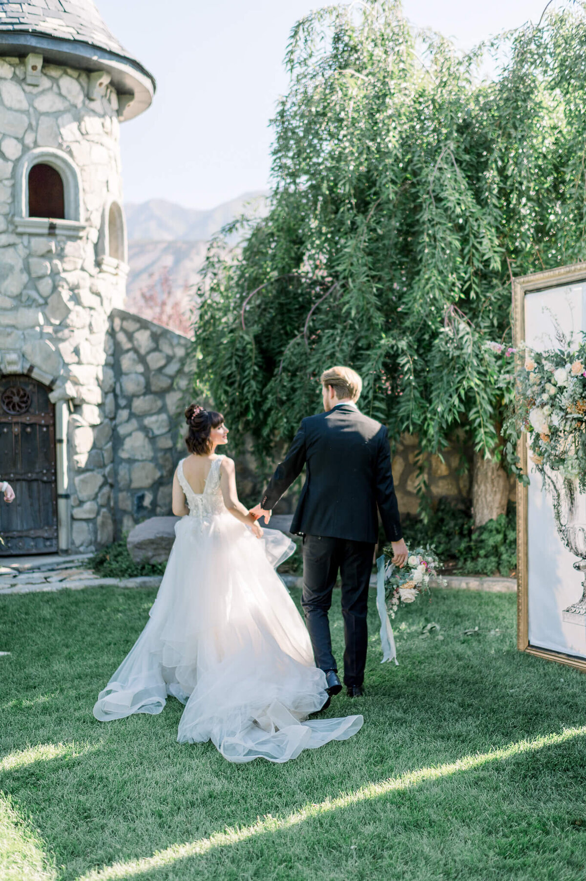 couple walks towards large castle wearing white dress and black tuxedo, groom holds wedding bouquet, arkansas wedding photographer