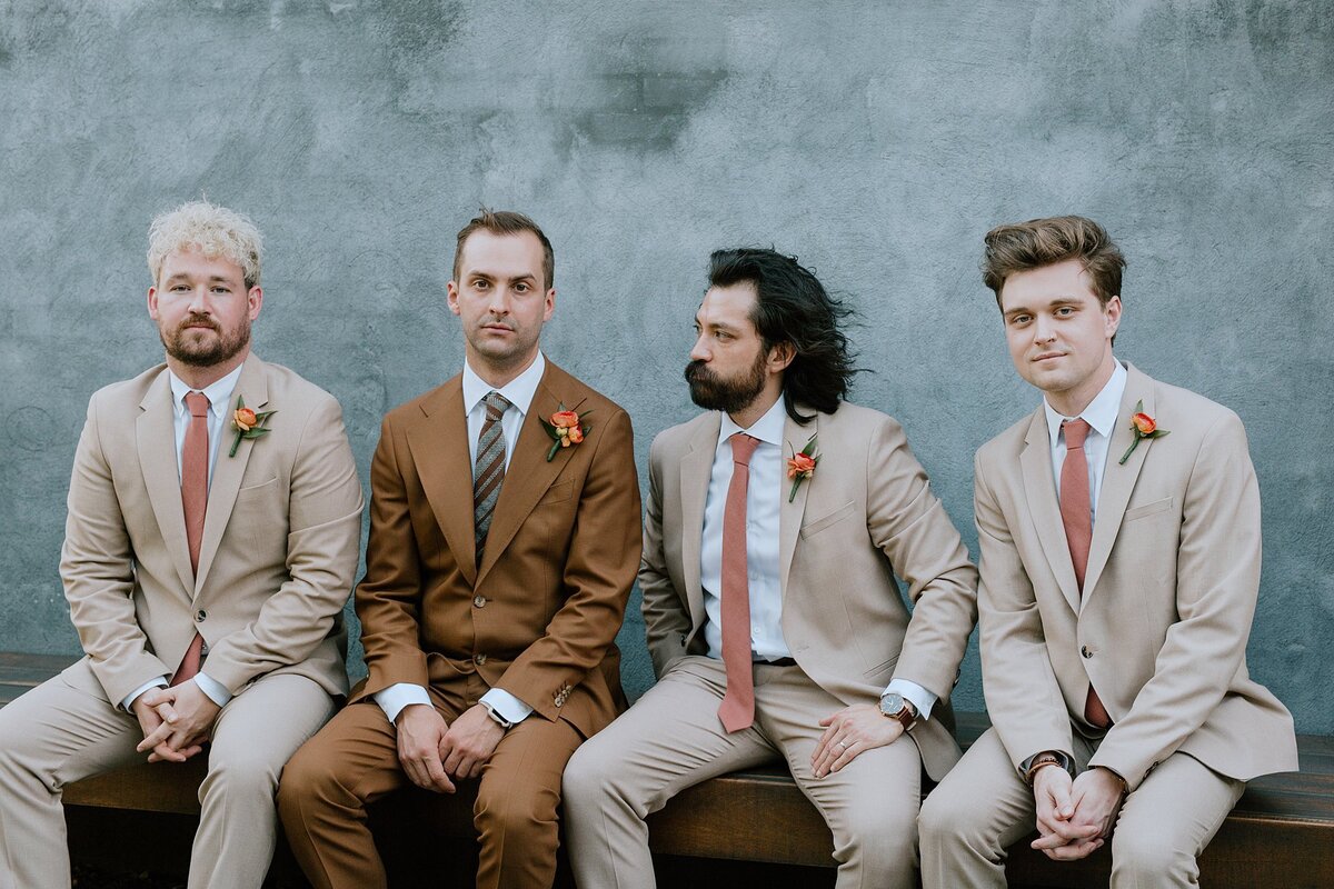 Nashville musicians at wedding. The groom, wearing a brown suit sits on a bench with his groomsmen and bandmates who are wearing tan suits with peach ties against a dusty blue wall  at Clementine Hall.