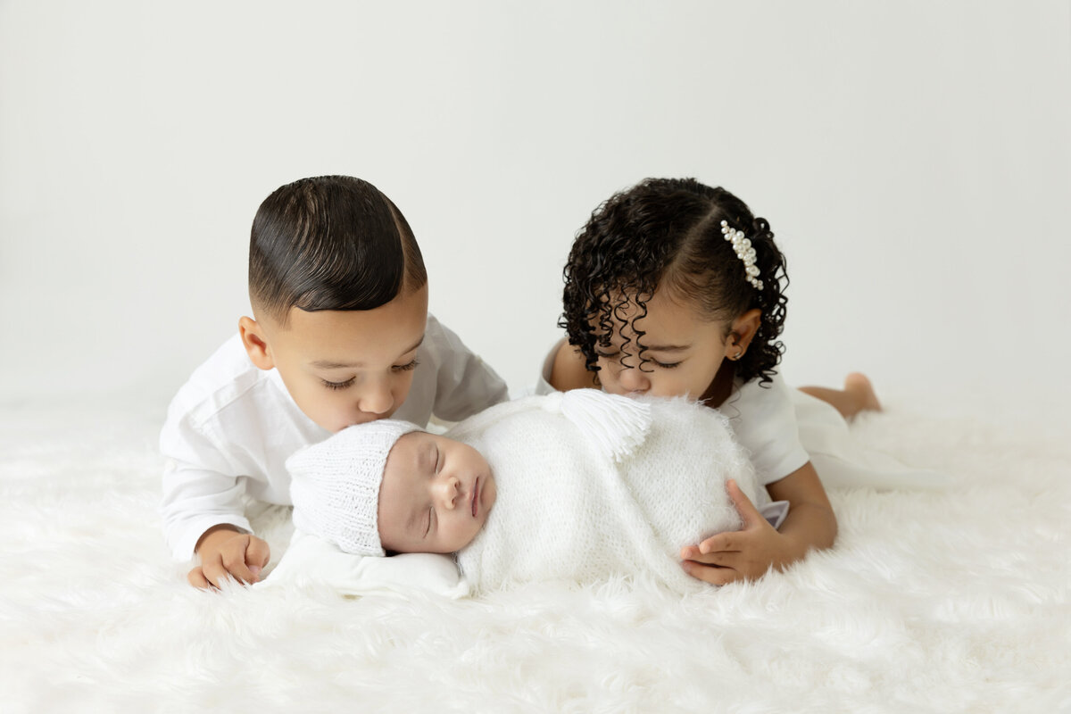 Toddler brother and sister kiss their sleeping newborn baby sibsling while laying on a white bed