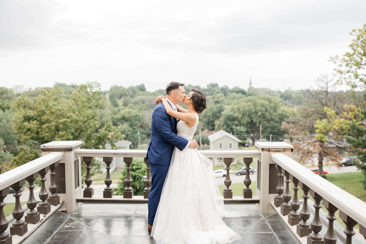 A newlywed couple embraces on a balcony overlooking a lush landscape, sharing a kiss, with the bride in a white gown and the groom in a blue suit, all beautifully orchestrated by their wedding coordinator from