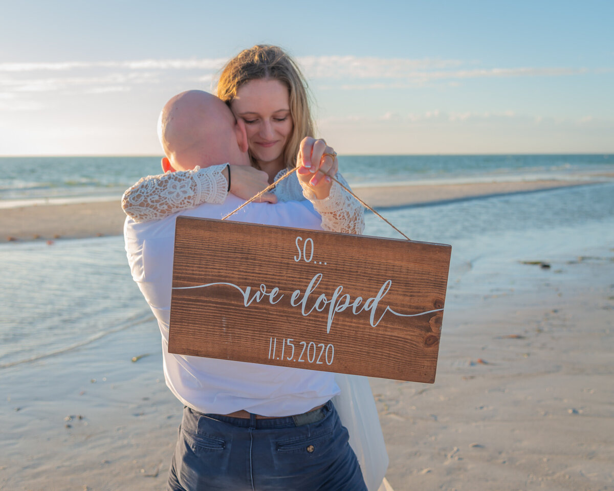 Groom holding his bride on the beach with a sign saying "So... We eloped"