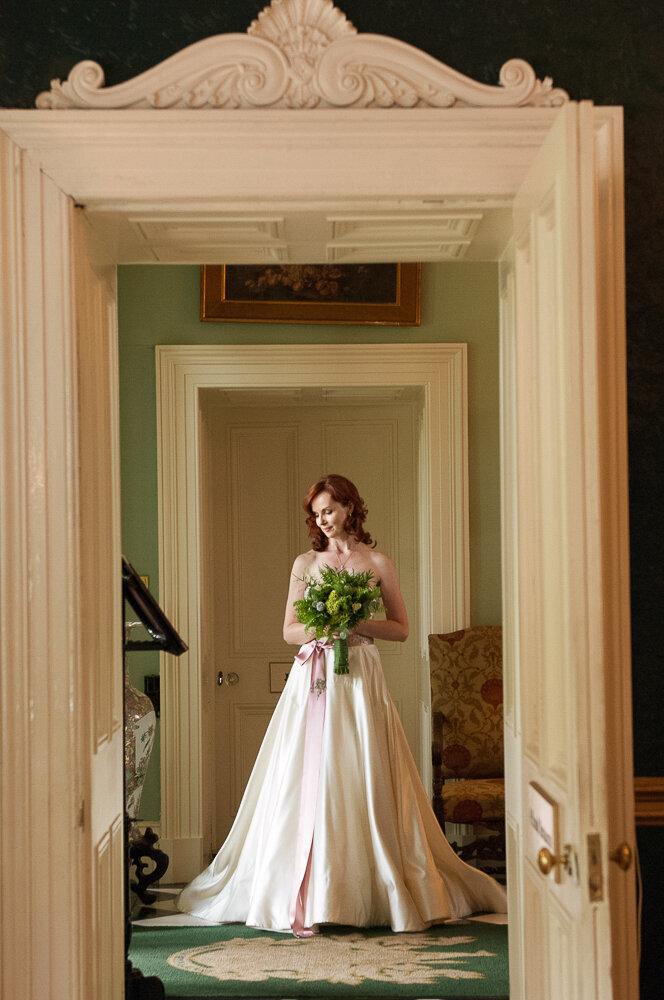 red-haired bride wearing an a-line style wedding dress with pink waistband and holding a green flower wedding bouquet while standing in the doorway of Ballyseede Castle
