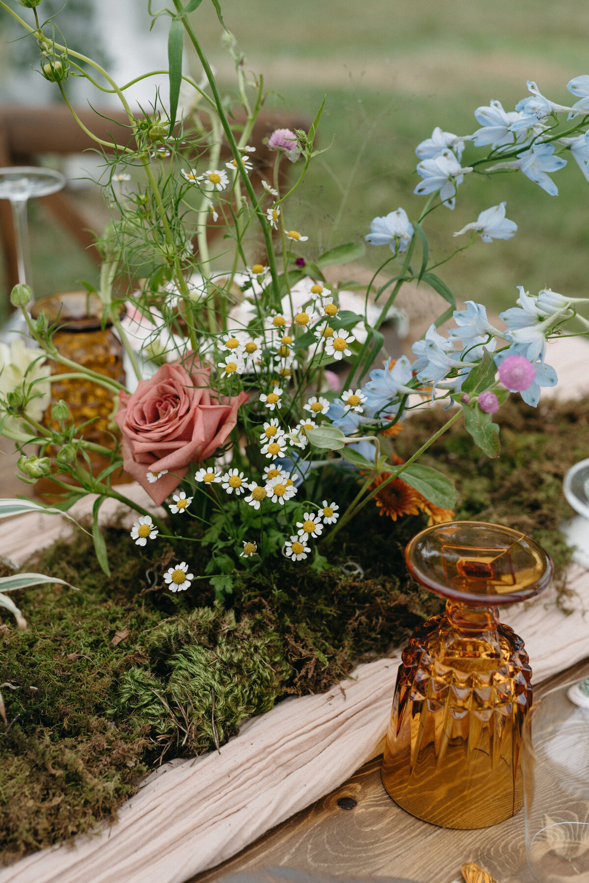 Close-up of a garden-inspired floral centerpiece with pink rose, blue delphinium, chamomile, and textured greenery arranged atop a natural moss runner for a whimsical outdoor wedding tablescape.