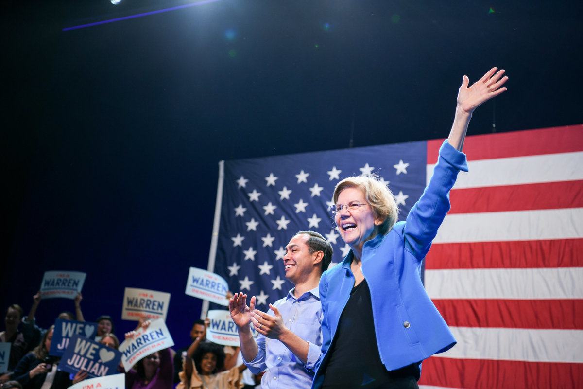 Elizabeth Warren and Julian Castro waving to the crowd in front of American flag