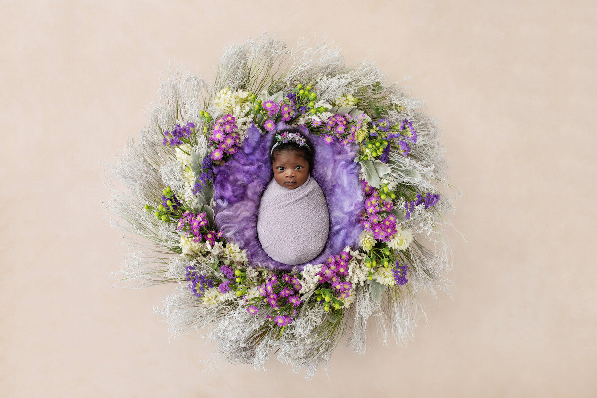 An awake newborn baby stairs up while laying in the middle of a floral wreath