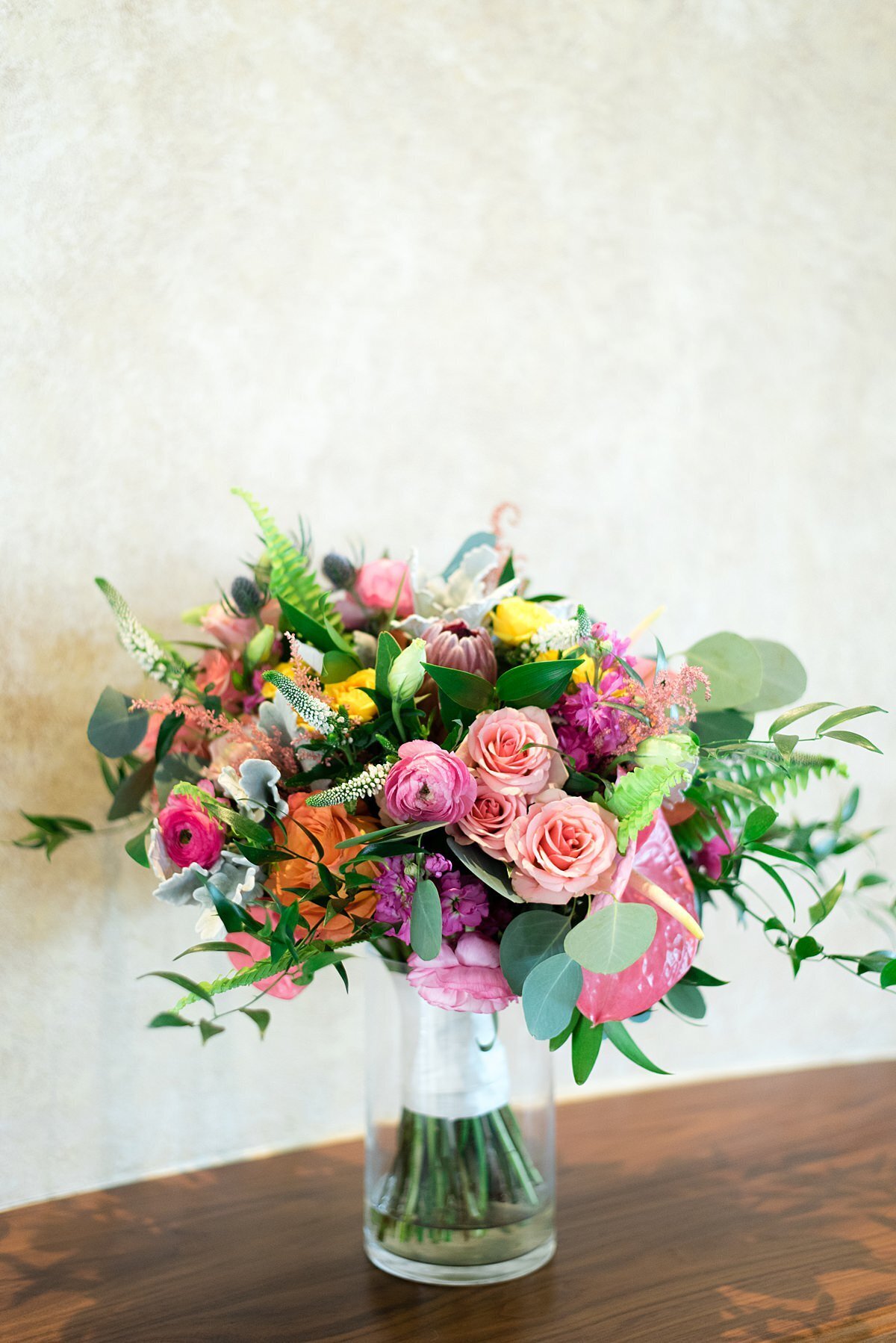A lush and colorful bridal bouquet wrapped with  white satin ribbon and is placed in a clear glass vase. There are  pink roses, pink ranunculus, orange roses, hot pink ranunculus, yellow roses, pink peonies, dusty miller, protea, king protea, and smilax vine in this very colorful bouquet.