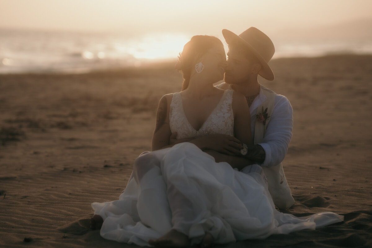 Mit dem Sonnenuntergang im Hintergrund sitzt das Hochzeitspaar in einer engen Umarmung hintereinander am Strand.